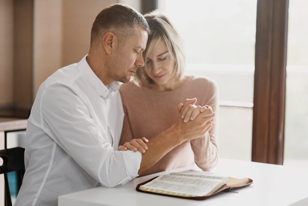 A husband and wife holding hands and praying together over an open Bible at a white table.