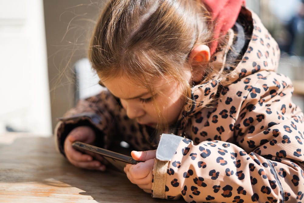 A little girl in a leopard-print coat bending over a smartphone and absorbed in its screen.
