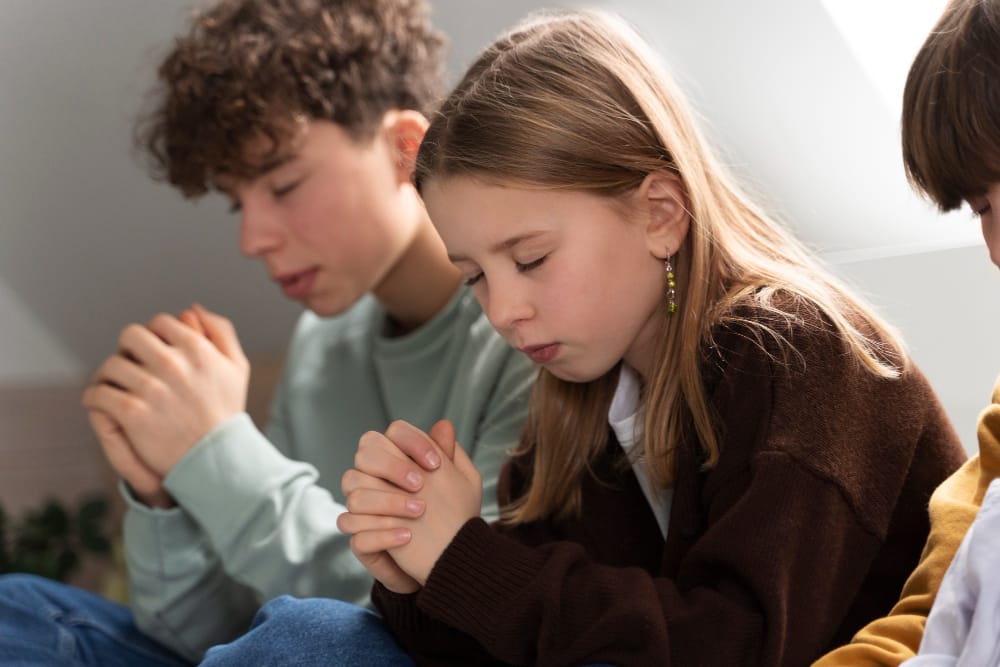 A boy and a girl sitting side by side with their eyes closed and hands folded in prayer.