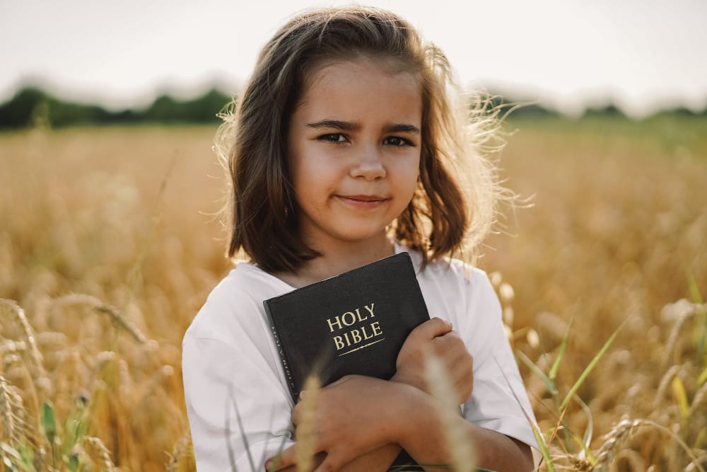 A young girl standing in a golden wheat field hugging a black Holy Bible to her chest.