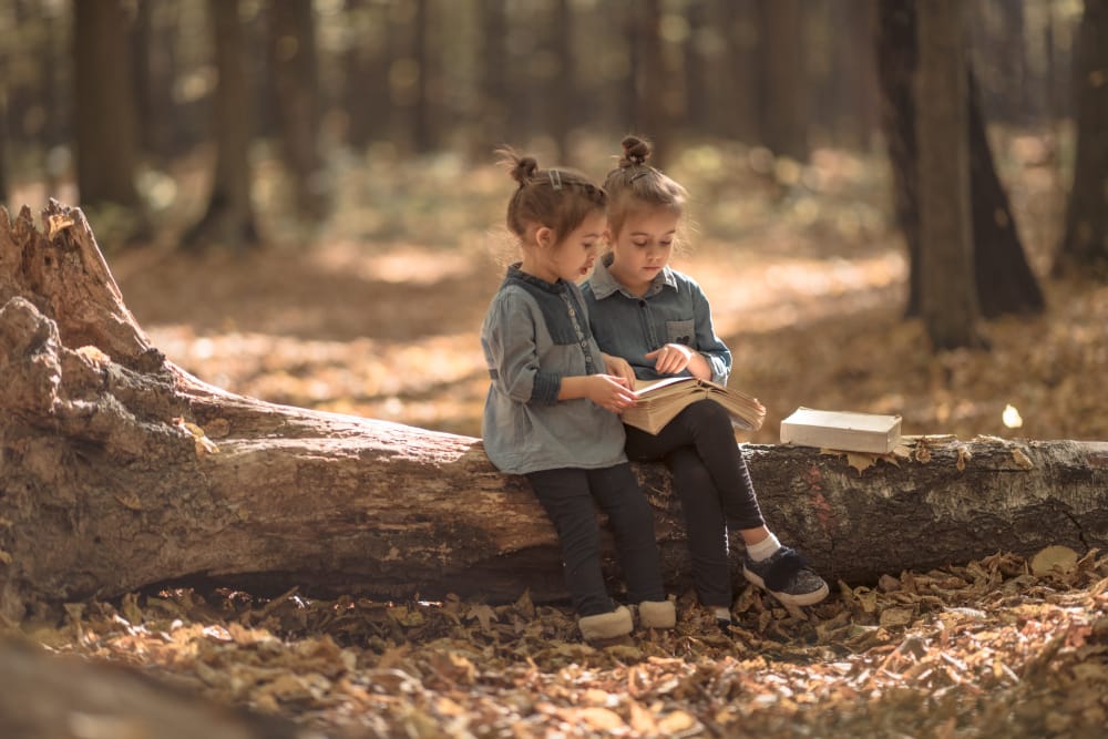 Two little girls sitting on a fallen log in an autumn forest, reading a book together.
