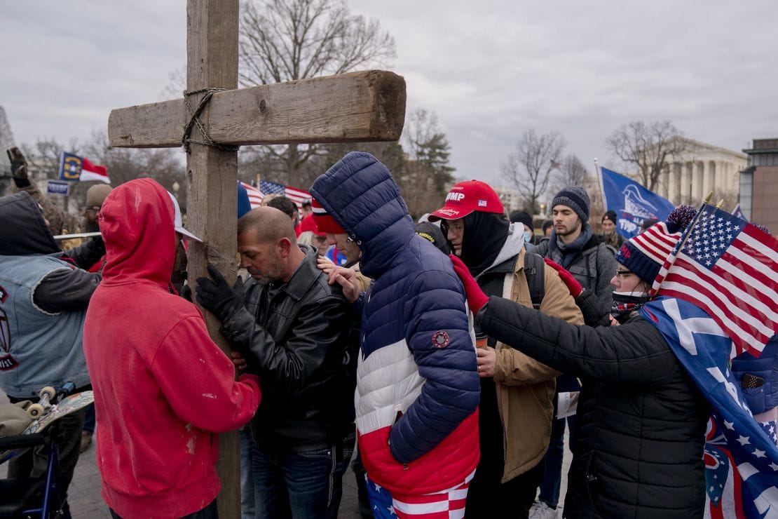 A crowd in winter coats and American flags gathered around a large wooden cross in front of a neoclassical government building.