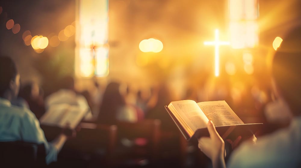 A worshipper holding an open Bible in a softly lit church, with a glowing cross shining in the blurred background.