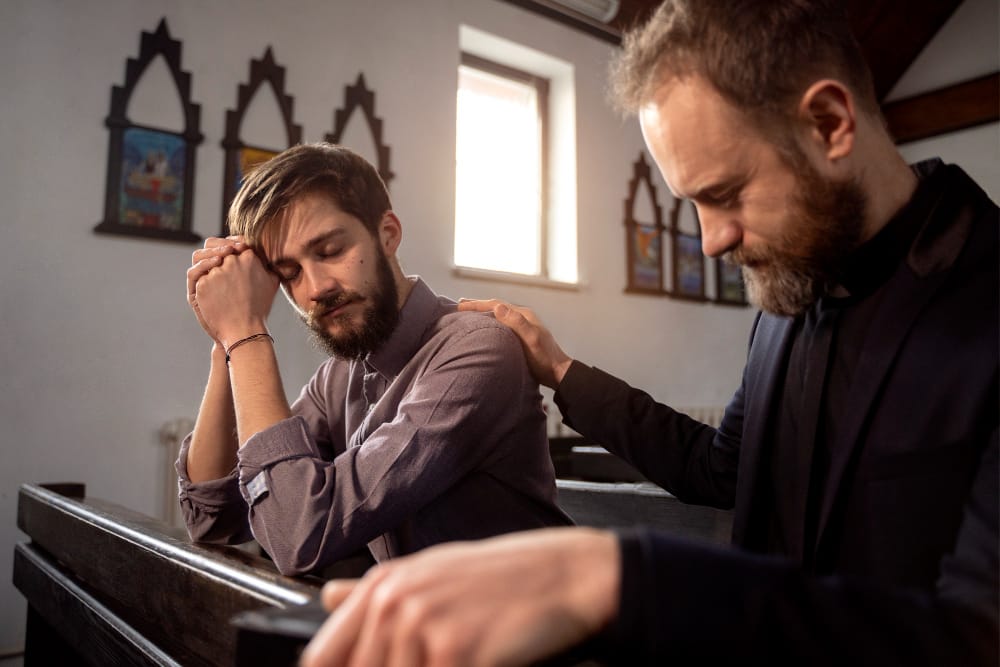A weary pastor in a pew with clasped hands against his forehead while another man rests a comforting hand on his shoulder.