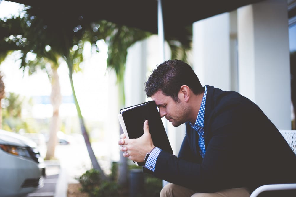 A pastor in a navy blazer sitting outdoors with his forehead pressed against a closed Bible, showing the weight of ministry burnout.