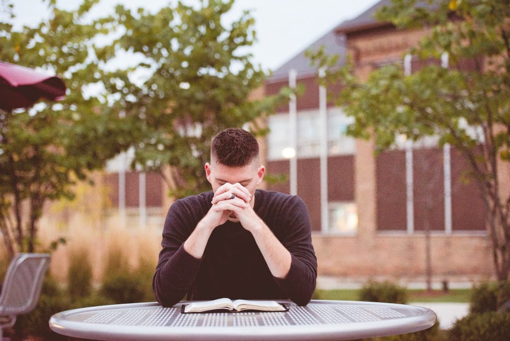 A man at an outdoor table with his head bowed and hands clasped over an open Bible.