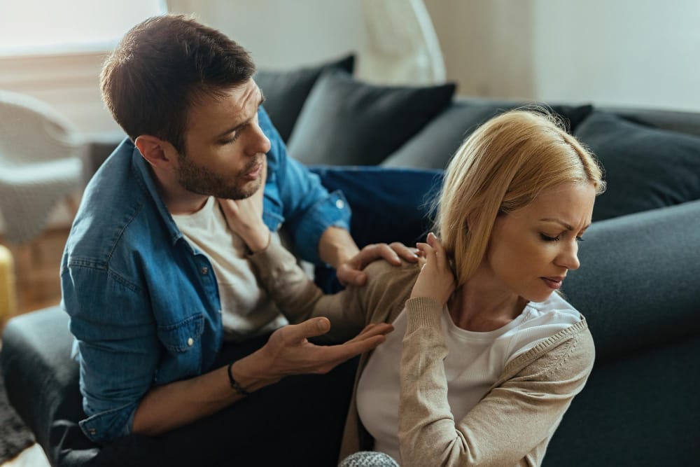 A man gesturing in appeal to his wife on a sofa while she turns away, upset, in a marriage divided by faith.