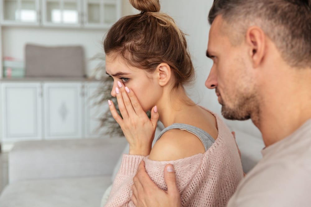 A woman wiping tears from her face on a sofa while her husband puts a hand on her arm to comfort her.
