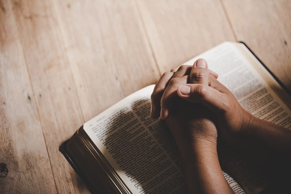 Clasped hands resting on an open Bible on a pale wooden table, seen from above.