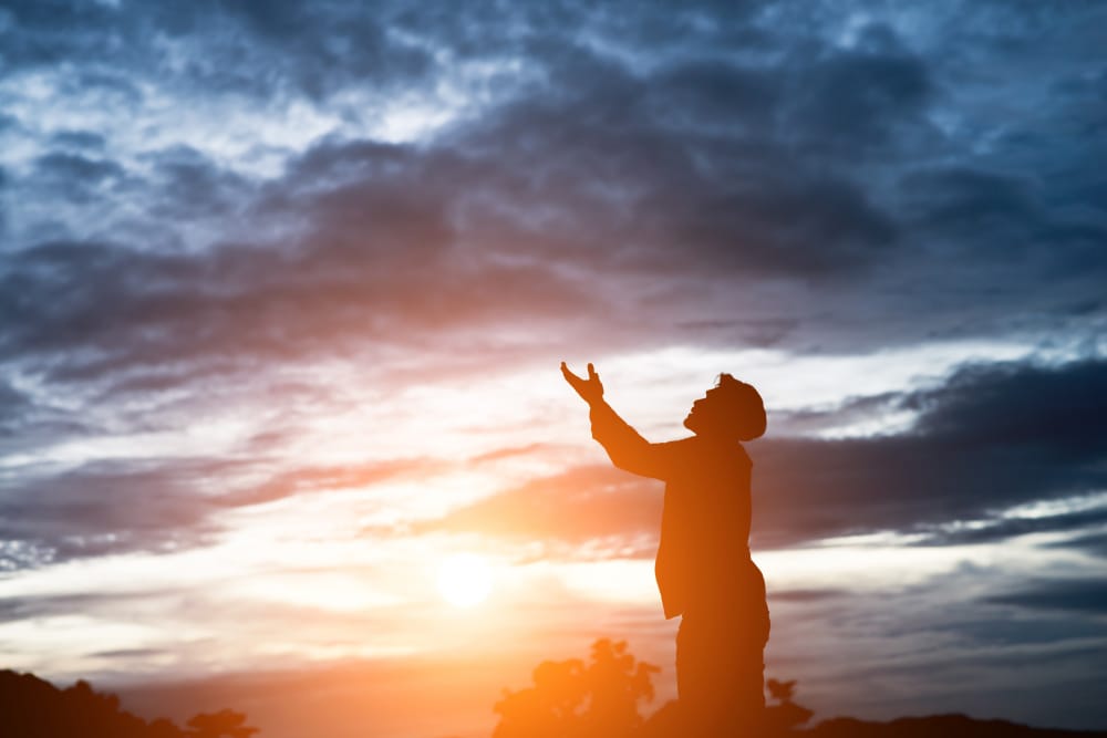 A silhouetted man raising his hands toward a dramatic sunset sky in prayer.