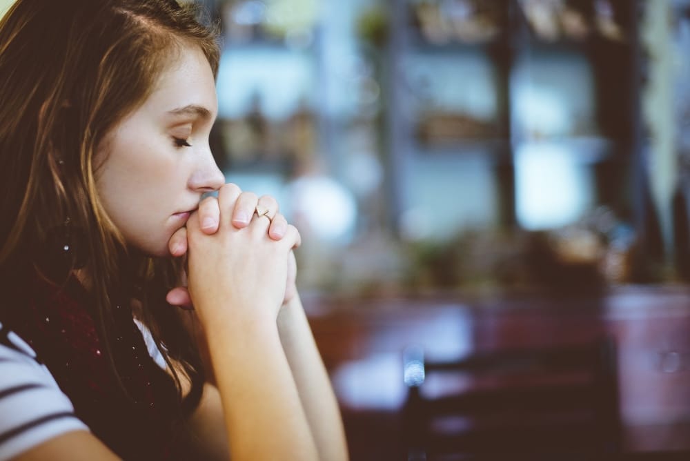 A young woman with her eyes closed and folded hands pressed to her lips in quiet prayer.