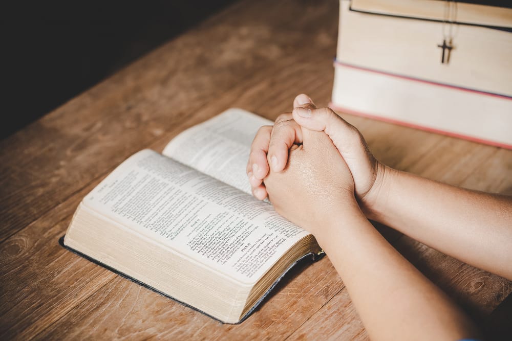 A person's clasped hands resting on an open Bible on a wooden table, with a stack of books and a cross necklace beside it.