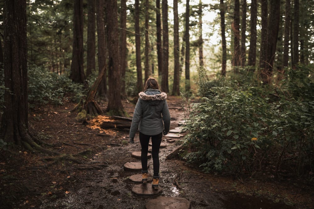A woman in a grey parka walking away along a muddy forest path of stepping stones between tall trees.