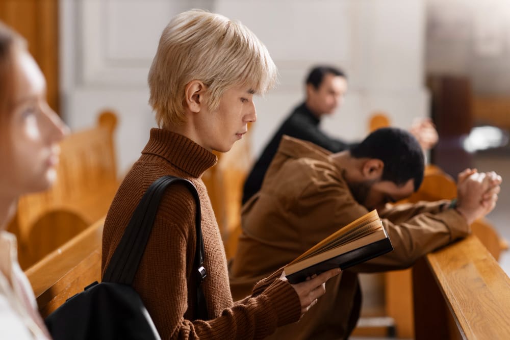 Worshippers sitting in wooden church pews, one reading a hymn book and another bowed over folded hands in prayer.