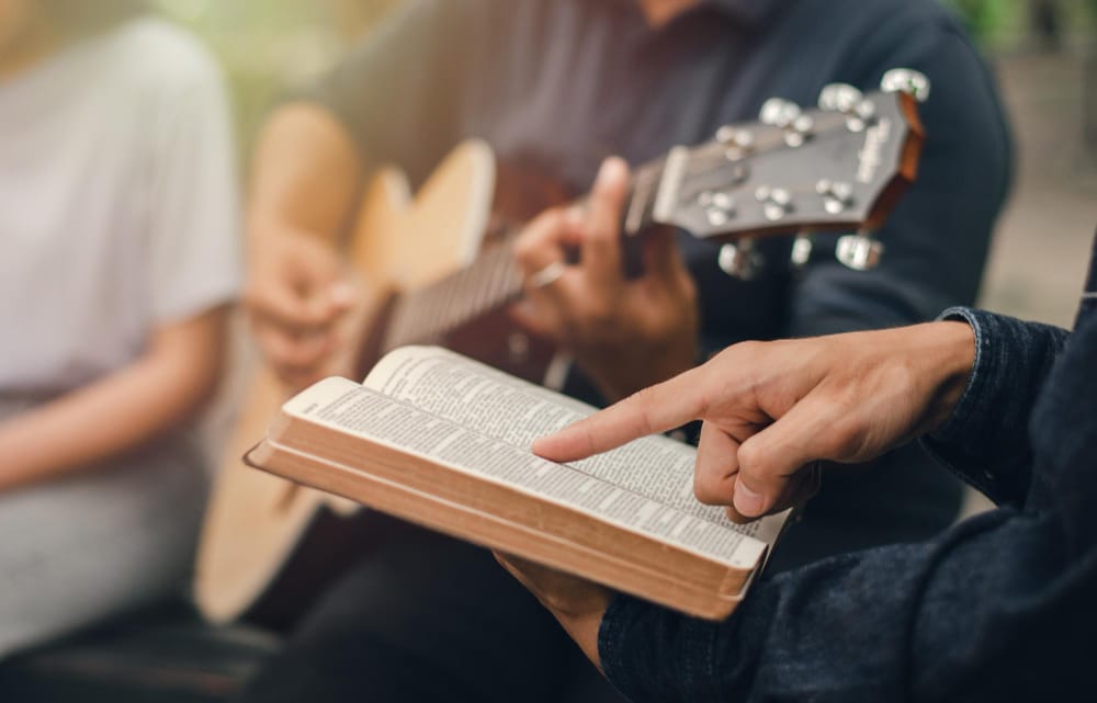 A small group with an acoustic guitar, one person pointing to a passage in an open Bible.