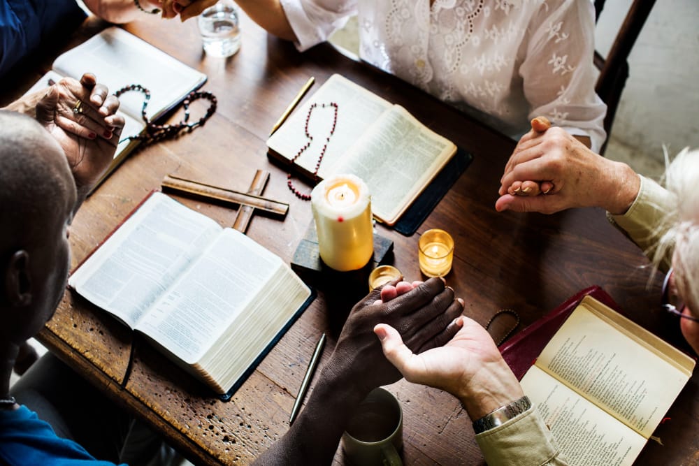 Overhead view of people holding hands in prayer around a wooden table with open Bibles, a rosary, a small cross and a lit candle.