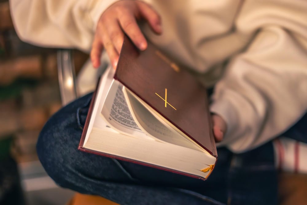 A person in a cream sweater holding a closed burgundy Bible with a gold cross on its cover on their lap.