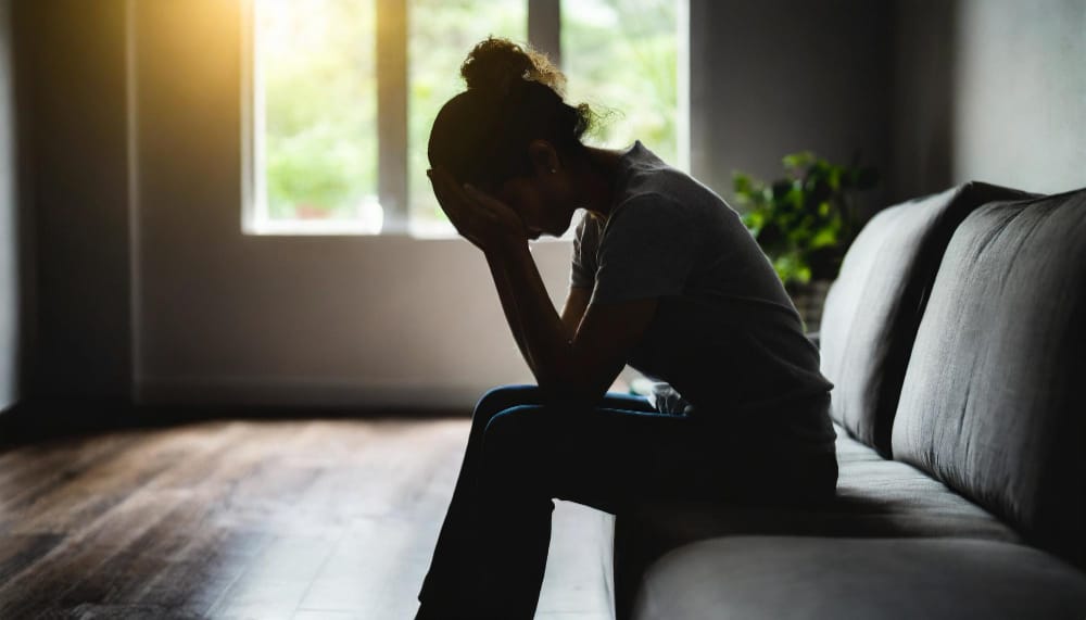 A woman in silhouette sitting on a sofa with her face in her hands in a quiet room.