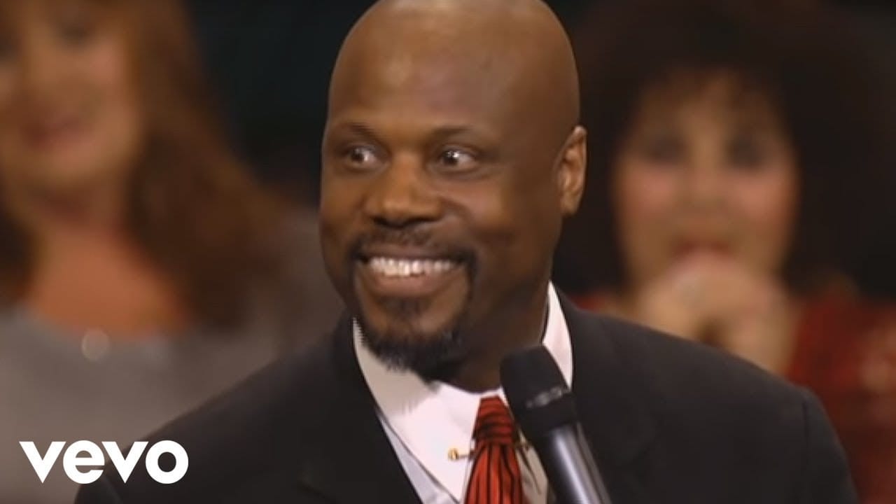 Pastor Wintley Phipps smiling in a dark suit and red tie as he sings into a microphone, in a still from a performance video.