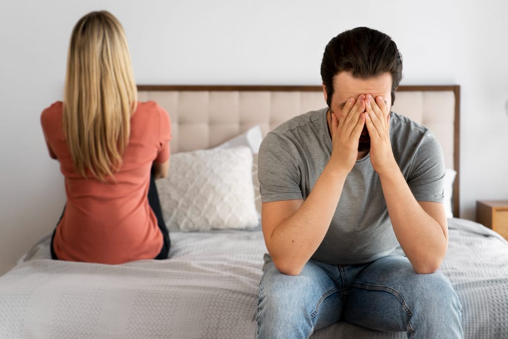 A woman sitting with her back turned on the edge of a bed while her partner beside her covers his face with his hands.