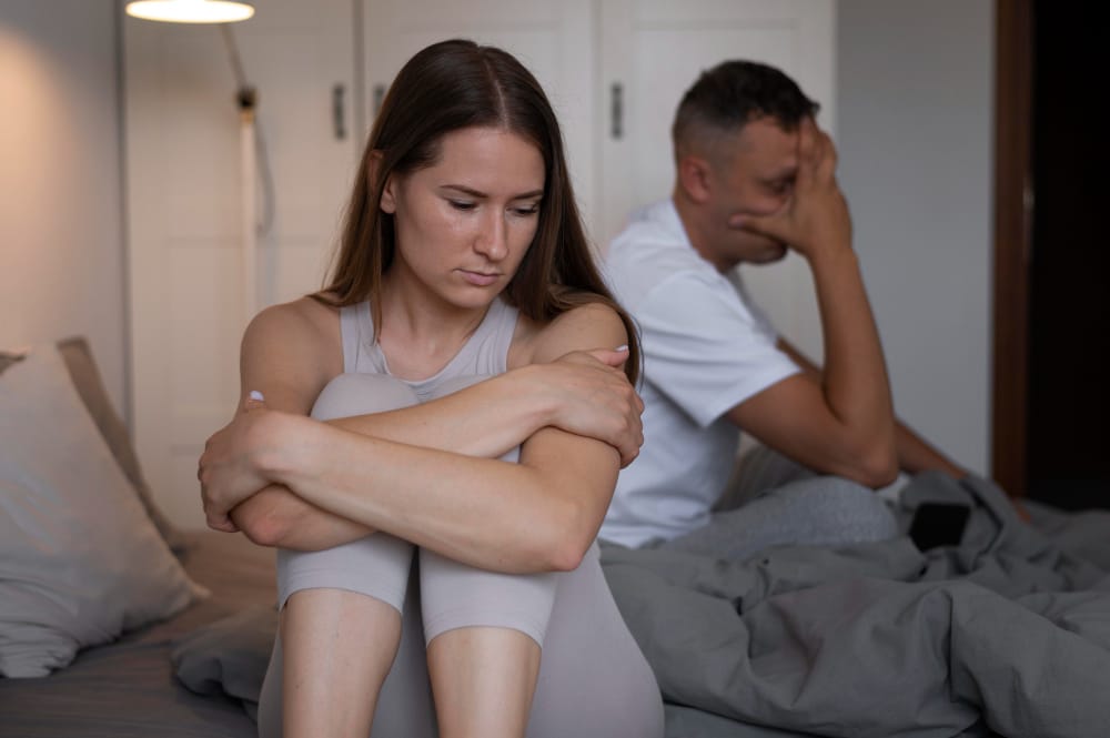 A woman hugging her knees and looking down while her partner sits behind her with a hand on his face on an unmade bed.