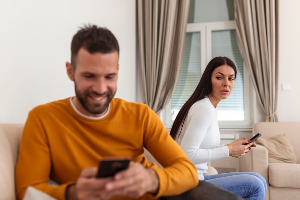 A man smiling at his phone on a sofa while his partner looks on with a suspicious expression.