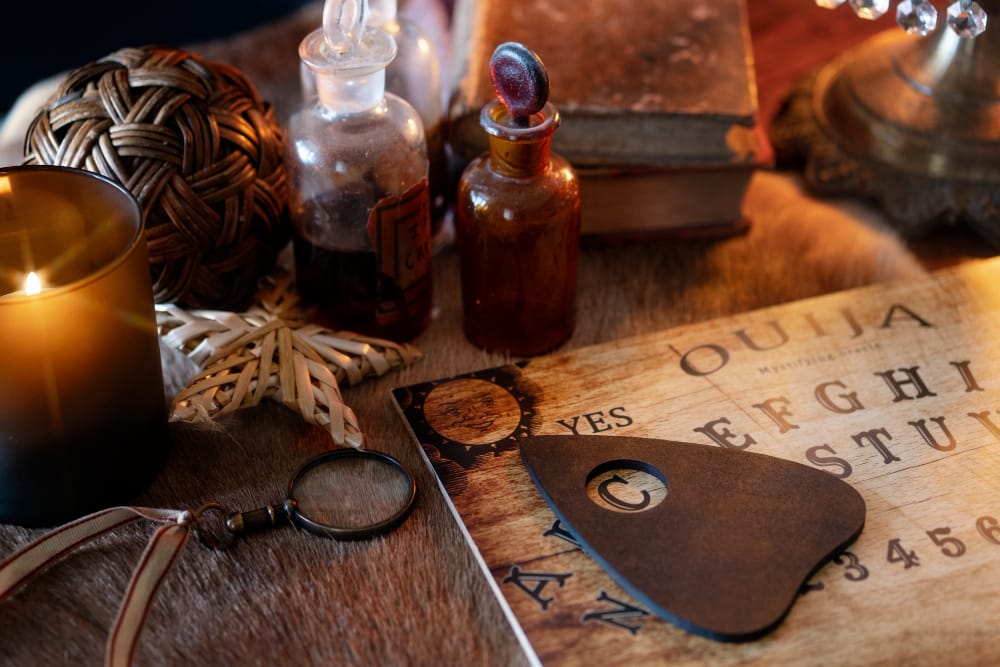A Ouija board with its planchette on a table beside a lit candle, glass potion bottles, a magnifying glass and an old book.