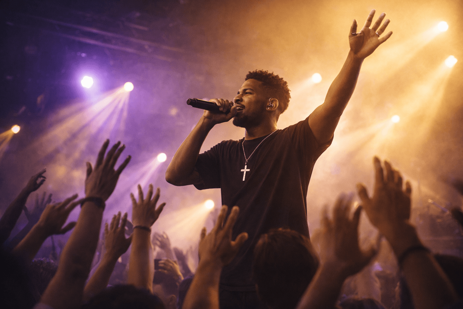 A singer wearing a cross necklace holding a microphone on a stage lit with purple spotlights, with a crowd's hands raised in front of him.