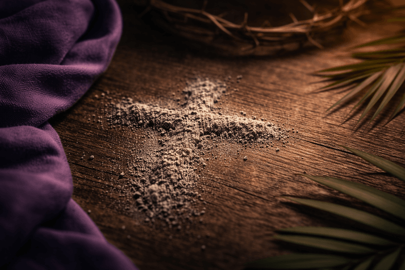A cross drawn in ashes on a wooden table beside purple cloth, palm leaves and a crown of thorns.