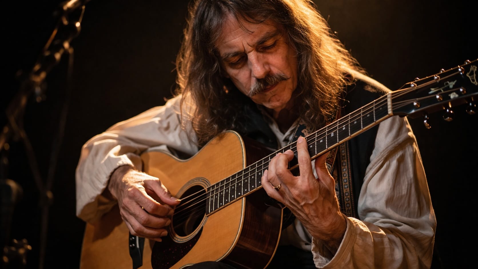 A long-haired, mustached guitarist playing an acoustic guitar in warm stage light.